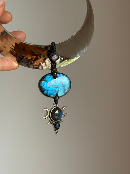 A handmade crescent moon pendant necklace held by a hand. The necklace features a hammered silver-toned crescent moon, a dark blue cord wrap, a round blue labradorite stone, and a dangling silver-toned triple moon and dark stone charm.
