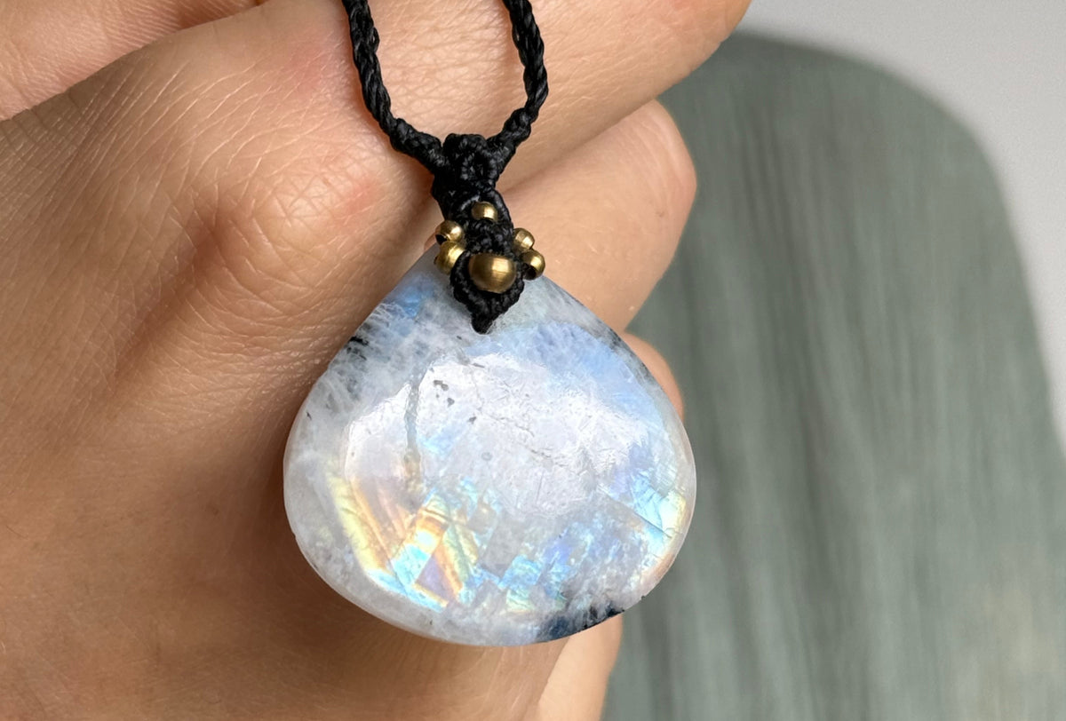 A hand holding a teardrop-shaped iridescent  moonstone pendant necklace with a black cord and small gold bead details.