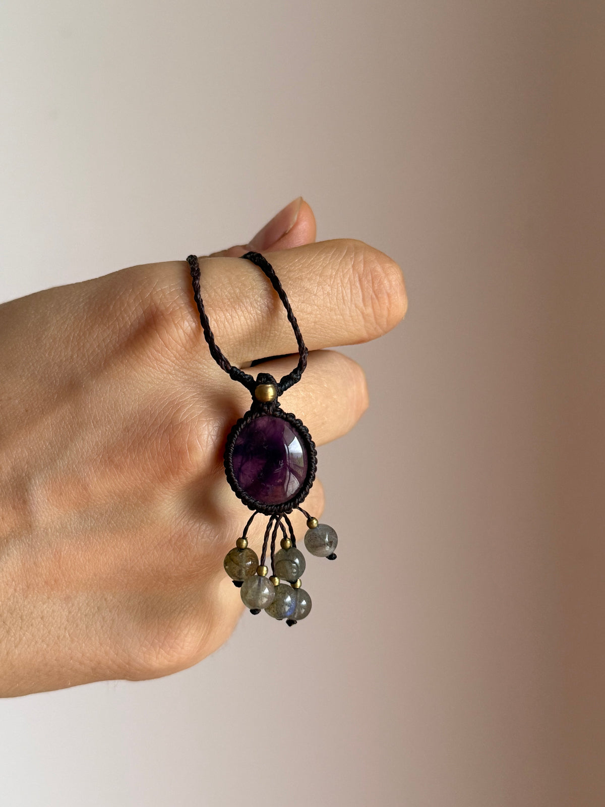 A hand holding a black cord pendant necklace with a oval amethyst and dangling labradorite beads, set against a plain beige background.