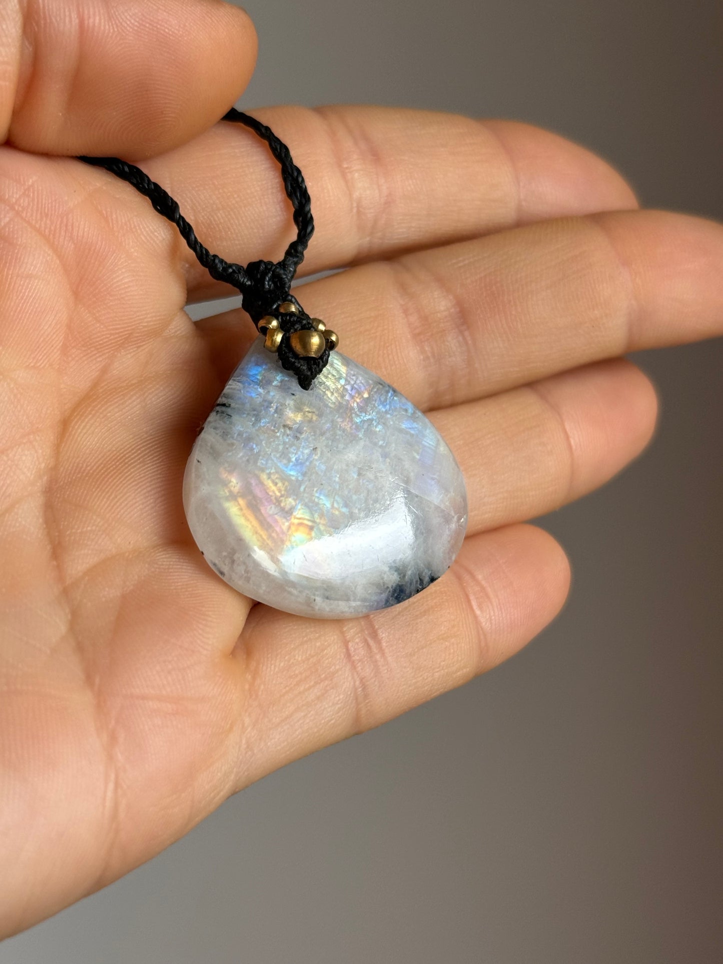 A teardrop-shaped iridescent moonstone pendant on a black braided cord, held in a person's hand against a neutral gray background.