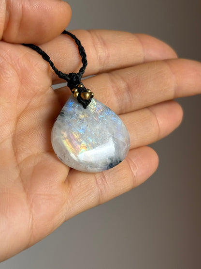 A teardrop-shaped iridescent moonstone pendant on a black braided cord, held in a person's hand against a neutral gray background.