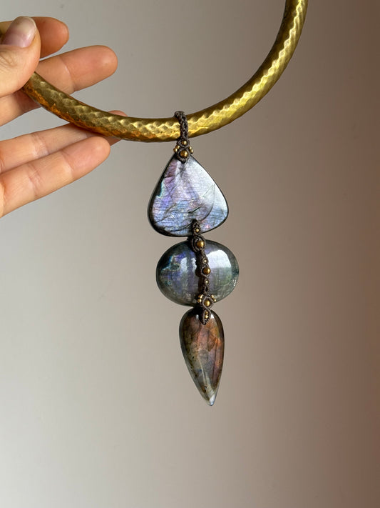 A close-up of a hand holding a chunky gold-tone necklace with three iridescent purple labradorite stones dangling from it, set against a plain beige background. with micro macrame details