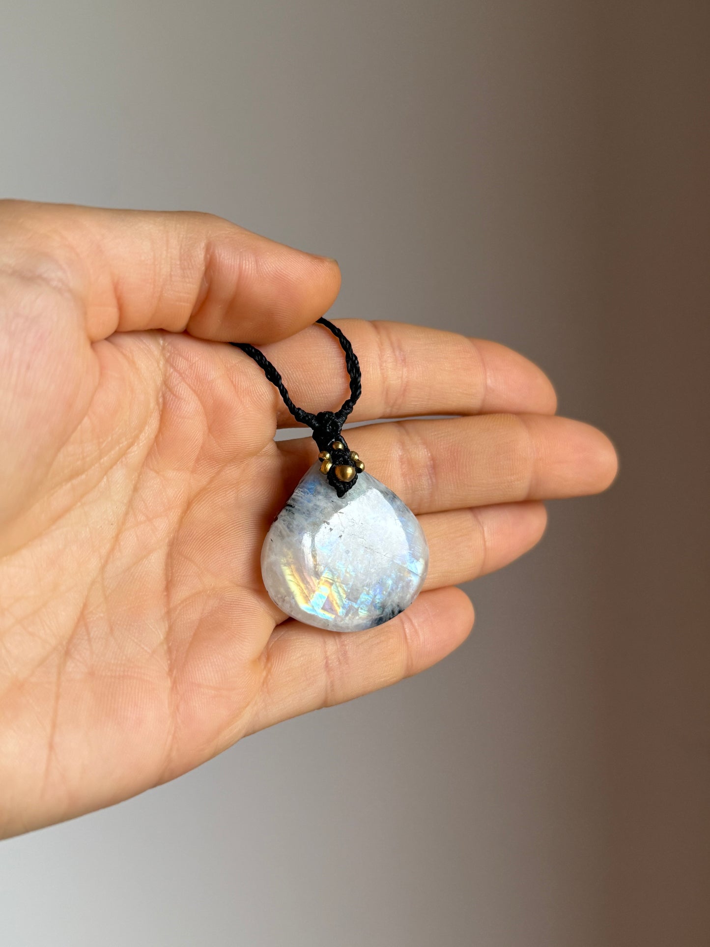 A hand holding a teardrop-shaped iridescent moonstone pendant necklace on a black cord. The pendant has a white and blue base with subtle rainbow flashes, and a small gold-toned accent at the top.