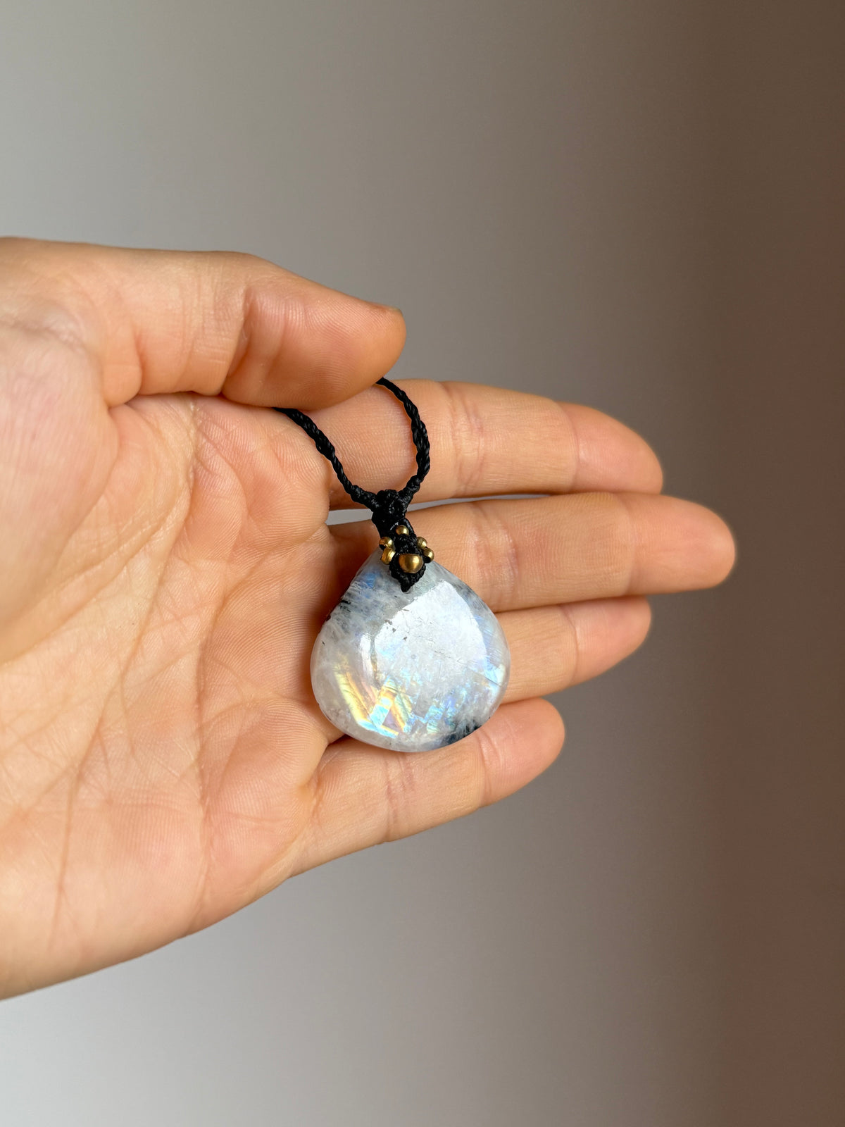 A hand holding a teardrop-shaped iridescent moonstone pendant necklace on a black cord. The pendant has a white and blue base with subtle rainbow flashes, and a small gold-toned accent at the top.