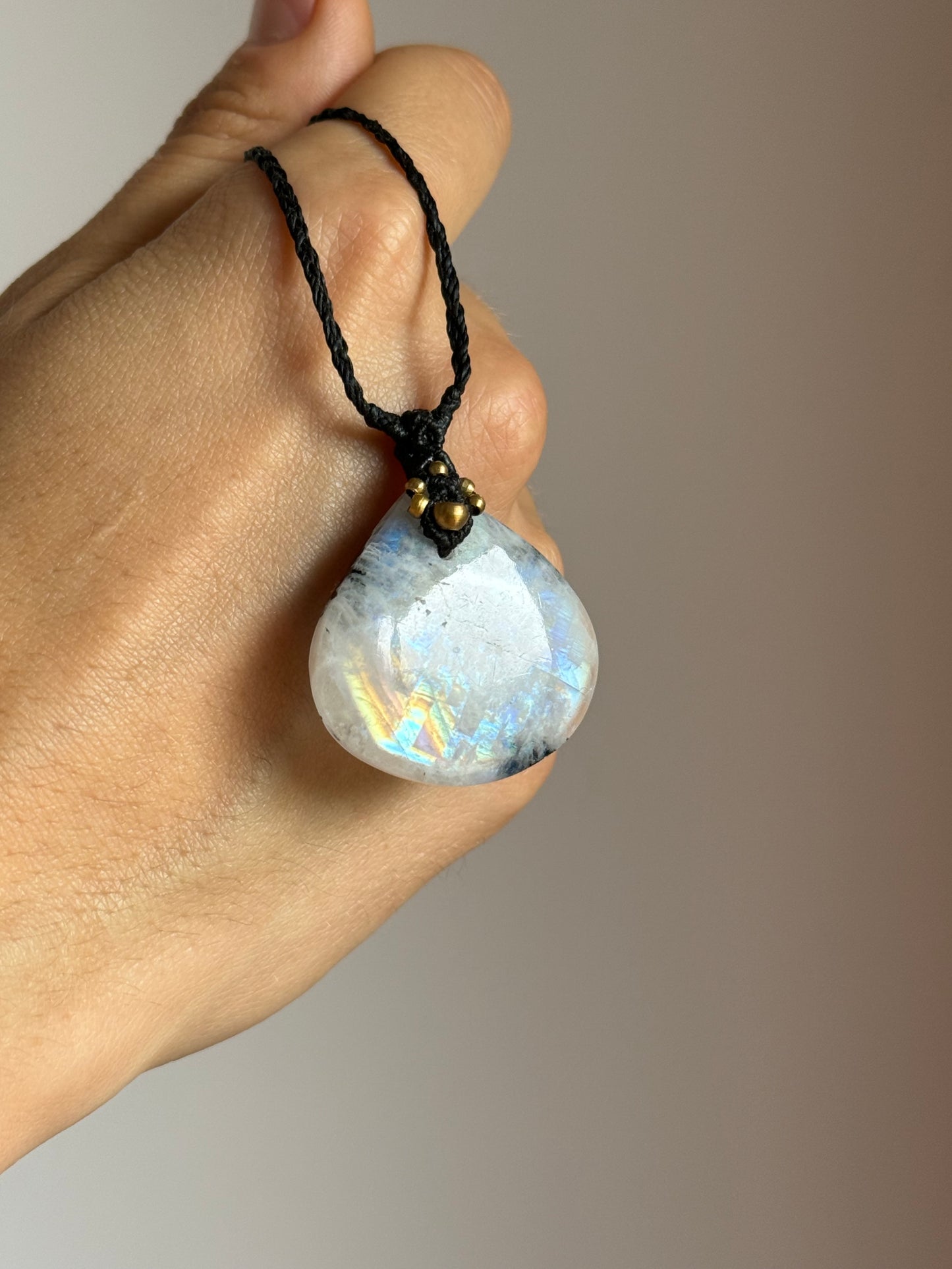 A hand holding a teardrop-shaped moonstone pendant on a black cord necklace, showing the iridescent play-of-color of the stone.