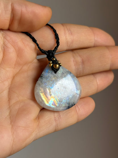 A teardrop-shaped moonstone pendant with iridescent blue and gold flashes, hanging from a black braided cord and held in a person's hand.